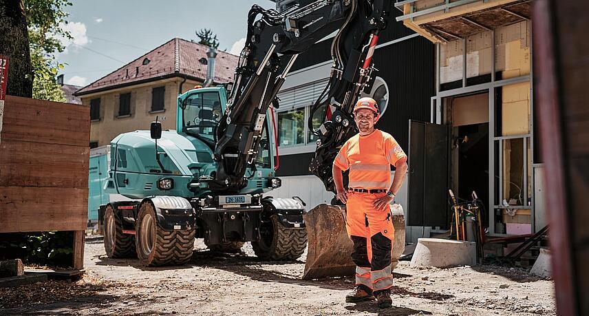 Operátor v pracovnom odeve s vysokou viditeľnosťou stojí vedľa kolesového rýpadla Wacker Neuson EW100 na stavebnom dvore v meste.