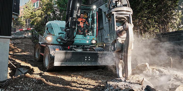 Kolesové rýpadlo Wacker Neuson EW100 s hydraulickým kladivom pri demolačných prácach v stiesnenej obytnej oblasti.