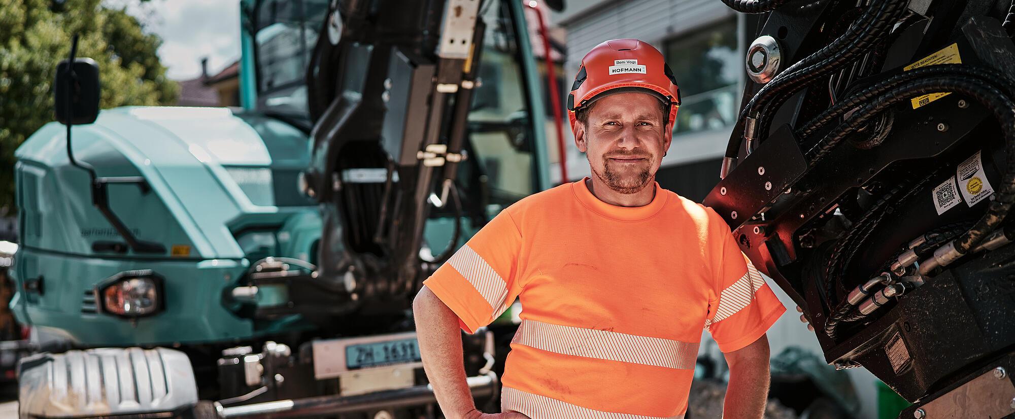Construction operator standing beside a Wacker Neuson EW100 wheeled excavator equipped with a hydraulic attachment.