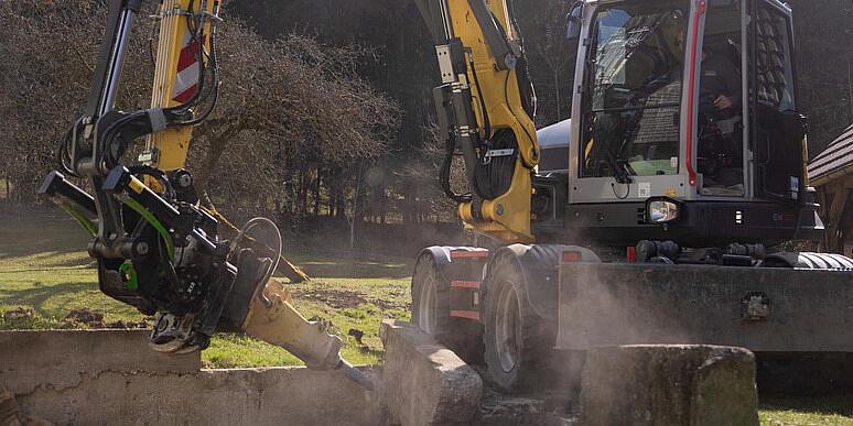 Kolesové rýpadlo Wacker Neuson EW100 obsluhujúce drvič pňov na lesnej čistinke, ktoré drví pň a vyhadzuje drevené štiepky do vzduchu, pričom v pozadí sú vidieť nahromadené polená a lesný porast.