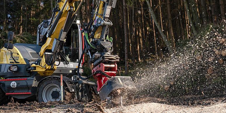 Kolesové rýpadlo Wacker Neuson EW100 obsluhujúce drvič pňov na lesnej čistinke, ktoré drví pň a vyhadzuje drevené štiepky do vzduchu, pričom v pozadí sú vidieť nahromadené polená a lesný porast.