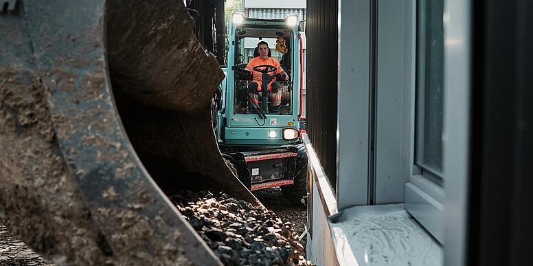 Bager Wacker Neuson EW100 prepravuje kamenivo lopatou v úzkej stavebnej zóne vedľa budovy.