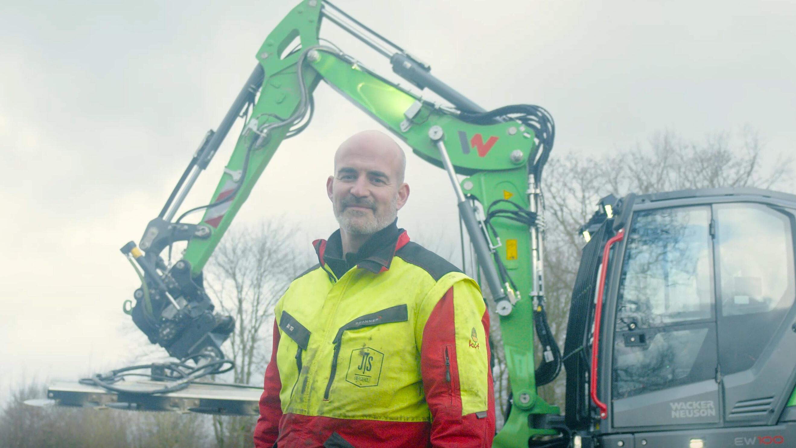 Operator standing in front of a Wacker Neuson EW100 wheeled excavator equipped with a tiltrotator attachment.
