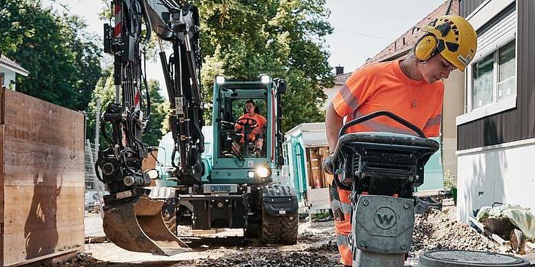 Stavebný robotník obsluhujúci vibračný zhutňovač značky Wacker Neuson, zatiaľ čo v pozadí pracuje rýpadlo EW100.