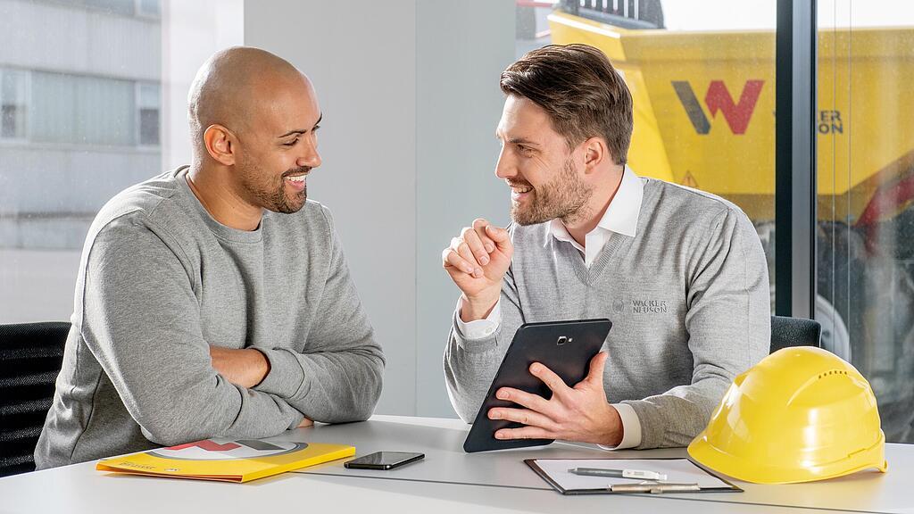 Zamestnanec Wacker Neuson sedí na písacom stole a ukazuje niečo zákazníkovi na tablete.