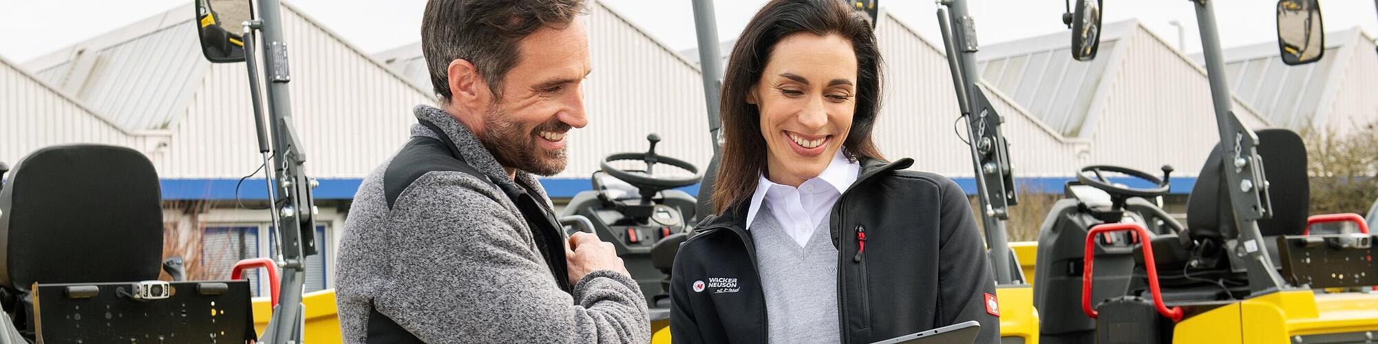 Usmiata zamestnankyňa Wacker Neuson s tabletom radí usmiatemu zákazníkovi pred stavebnými strojmi Wacker Neuson.